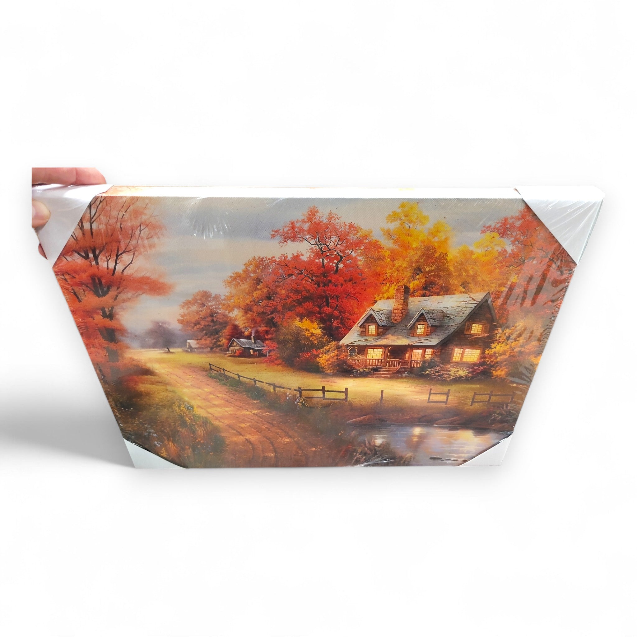 Eine Hand hält das LED-Wandbild "Herbstwald" von Weltbild (40x30 cm), das ein Häuschen an einer Straße und einem Teich inmitten leuchtender Herbstbäume zeigt - eine ideale herbstliche Wanddekoration für jeden Raum.