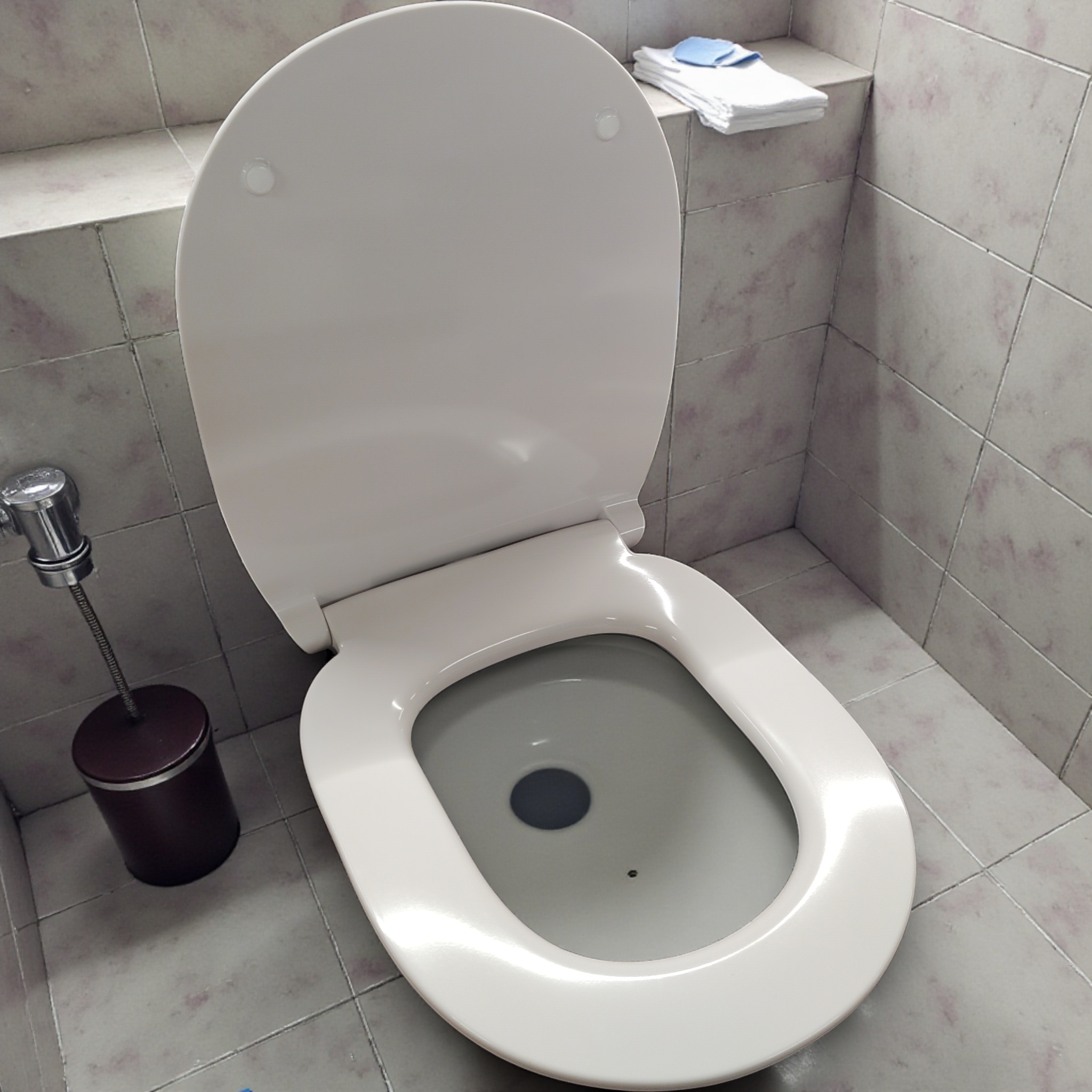 Offene Toilette mit weißem ultraflachen WC-Sitz mit Absenkautomatik von Ideal Standard Connect in einem gefliesten Badezimmer; linksseitiges Spülventil und Ablage mit gefalteten Tüchern sichtbar. Kompatibilität: E772401 enthalten.