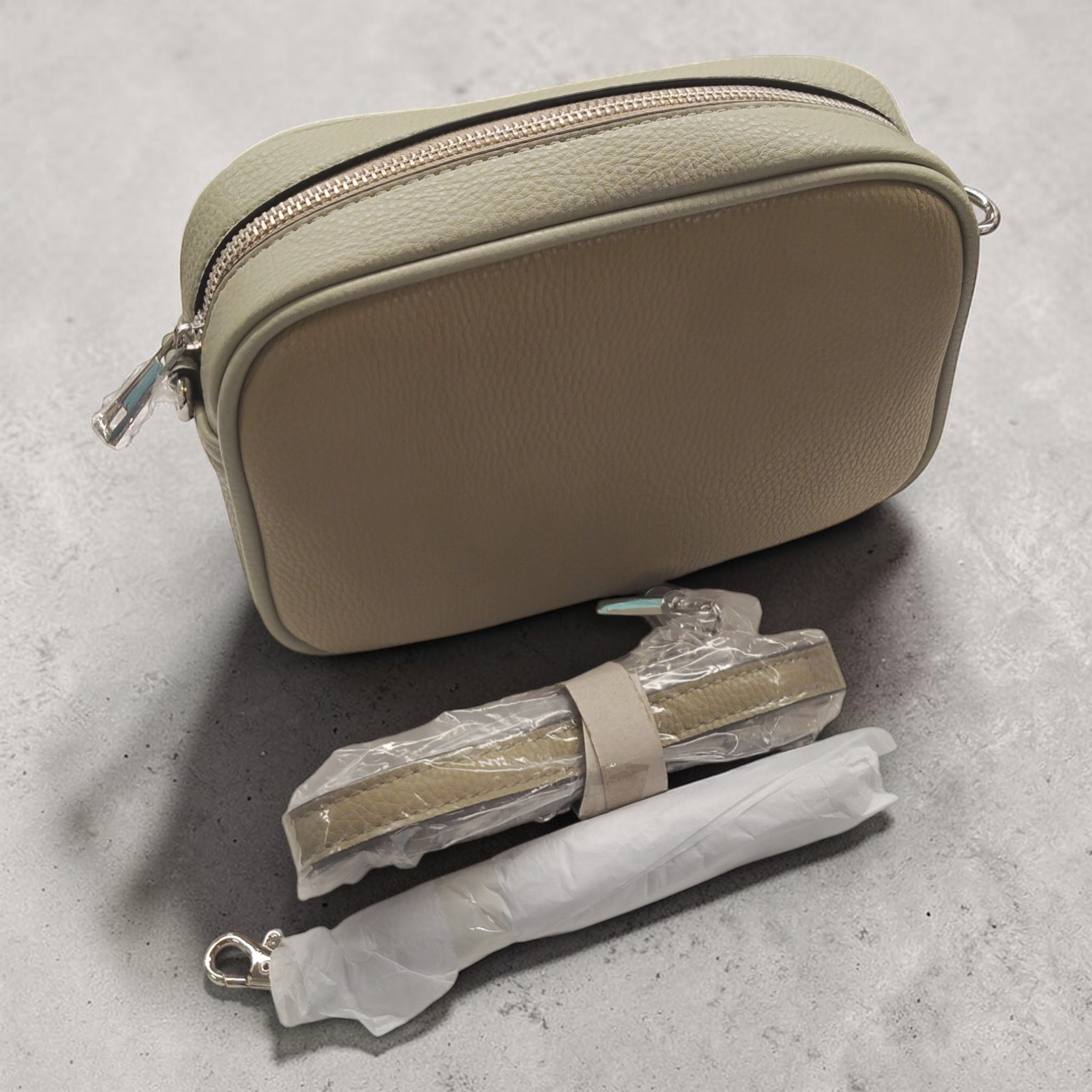 Eine kleine beige Schultertasche Umhängetasche von Collezione Alessandro, neu und Made in Italy, mit Reißverschluss und zwei abnehmbaren, in Schutzfolie verpackten Trägern liegt auf einer grauen Fläche.