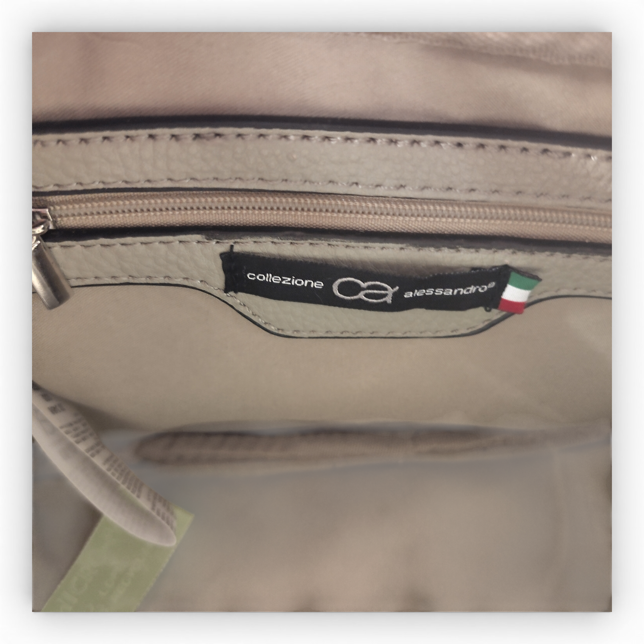 Nahaufnahme der Innenseite einer beigen Schultertasche Umhängetasche von Collezione Alessandro, mit einer Reißverschlusstasche und einem schwarzen Etikett mit dem Markennamen und einer italienischen Flagge, das die Qualität "Made in Italy" hervorhebt.