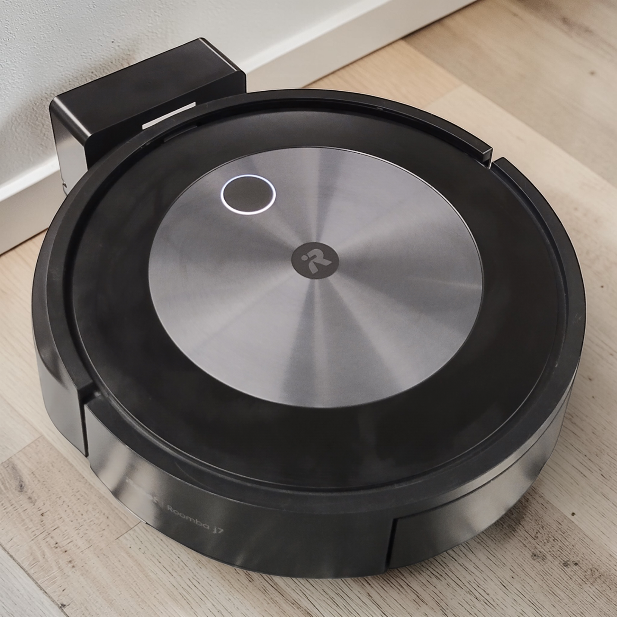 Ein iRobot Roomba J7+, Mod. RVE-Y1, / TOP gebraucht, mit Ein-Knopf Bedienung und App-Steuerung von iRobot steht auf einem Holzboden neben seiner Ladestation.